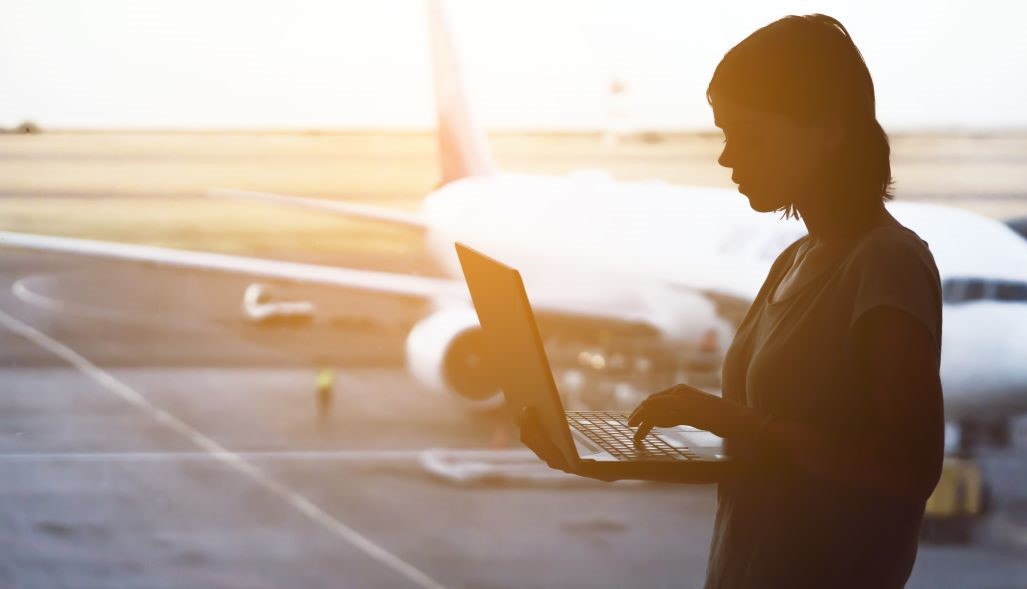 female holding laptop at an airport with a plane on the runway in the background